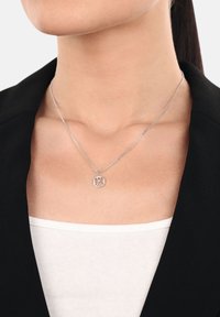 Collana d'argento con un pendente circolare che presenta figure intrecciate, su una delicata catena. Indossata su una maglia bianca, con un blazer nero.