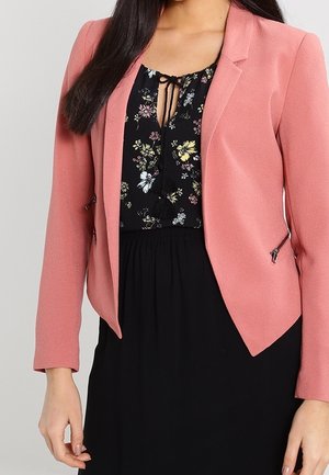 Vrouw met een roze blazer over een zwarte bloemenblouse en een zwarte rok, getoond van schouders tot halverwege de dij, staand tegen een effen achtergrond.