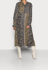 Długa, wzorzysta sukienka z printem zwierzęcym w odcieniach czerni, brązu i oliwkowego, z guzikiem przy dekolcie i marszczonymi mankietami, zestawiona z białymi botkami.