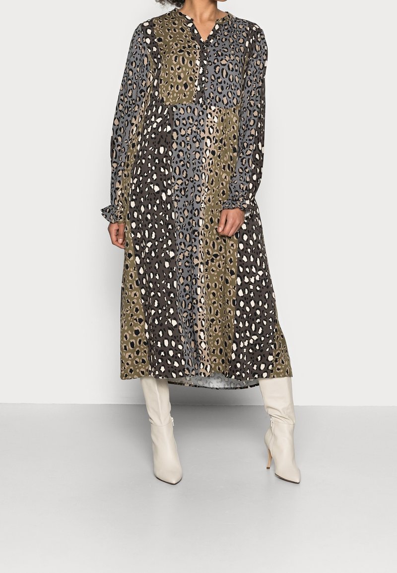 Długa, wzorzysta sukienka z printem zwierzęcym w odcieniach czerni, brązu i oliwkowego, z guzikiem przy dekolcie i marszczonymi mankietami, zestawiona z białymi botkami.