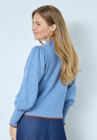 Világoskék pulóver bordázott textúrával, puffadt ujjal és ellentétes barna díszítéssel az alján. Denim szoknyával van stílusosan párosítva, semleges háttér előtt.