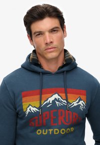 Felpa blu con coulisse, caratterizzata da una grafica montana e la scritta "SUPERDRY OUTDOOR" in rosso e giallo. Sotto c'è un motivo a quadri.