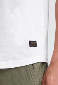 Hvid bomulds T-shirt med en sort label med guld tekst. Kanten har en lige skæring med synlige sting. Sammen med olivengrønne bukser.