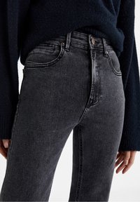 Jeans en denim noir à taille haute, présentant un léger délavage, un design classique à cinq poches et des accents en métal couleur cuivrée.