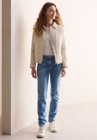 Chaqueta acolchada de color beige claro con cuello alto, camiseta blanca ajustada y jeans ajustados de color azul claro, combinados con zapatillas blancas.