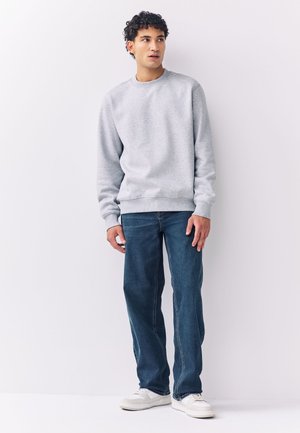 Mladý muž stojí, má na sobě světle šedou mikinu, modré džíny a bílé tenisky, mírně se dívá stranou na bílém pozadí.