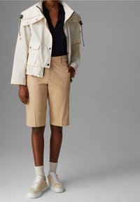 Giacca impermeabile bianca con colletto alto e cordoncini, maglietta blu navy, pantaloni shorts beige e sneakers crema con suole testurizzate.