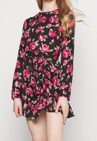 Musta kukkamekko, jossa on vaaleanpunaisia ruusuja, pitkät hihat, kiristetty vyötärö yhteensopivalla soljella sekä kerroksellinen hihansuu.