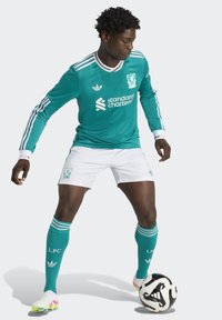 Maillot de football turquoise à manches longues avec un short blanc, présentant des accents rayés et le logo de Liverpool FC. Le joueur interagit avec un ballon noir et blanc.
