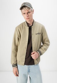 Khaki varsity jacket with dark brown accents, featuring snap buttons, striped cuffs, and a logo on the chest; light blue jeans.