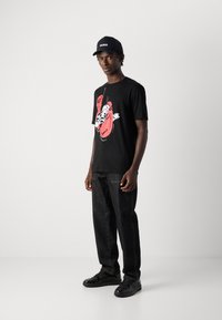 Bărbat îmbrăcat cu o șapcă neagră cu însemnul "HUGO," un tricou negru cu imprimeu cu mănuși de box roșii, blugi închiși la culoare și adidași negri, stând pe un fundal alb.