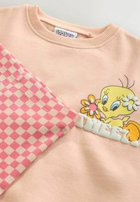 Lyserød sweatshirt med en tegneseriefigur, der holder en blomst. Venstre ærme har et lyserødt og cremefarvet ternet mønster med teksturerede accenter.