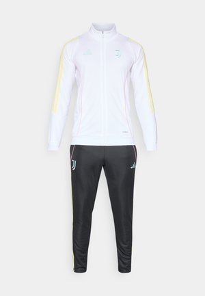 Tuta bianca con accenti gialli e rosa. La giacca presenta una chiusura con zip e un colletto alto. Pantaloni neri con loghi e strisce coordinate.