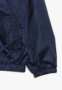 Giacca leggera blu navy con polsini elasticizzati, dotata di tasca laterale e texture liscia e lucida. Design semplice e finitura non decorata.