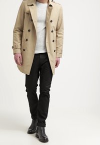 Trench coat beige con parte anteriore doppiopetto, bottoni neri ed epaulettes, abbinato a un maglione grigio, jeans neri e stivaletti neri.