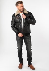 Schwarze Leder-Bomberjacke mit einem beigen Lammfellkragen, Zip-Taschen und Patches, kombiniert mit dunklen Jeans und braunen Lederstiefeln.