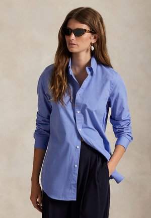 Une femme pose en portant une chemise bleue boutonnée, un pantalon foncé, des lunettes de soleil et des boucles d’oreilles pendantes, sur un fond beige uni.