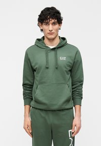 Zelená bavlnená mikina s kapucňou, predným vreckom, nastaviteľným sťahovacím šnúrkou a logom EA7 na hrudi. Dopĺňa ju zelené tepláky v rovnakom odtieni.