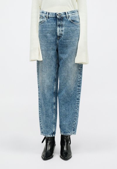 Jean en denim bleu clair à taille haute, avec une coupe ample et un délavage, présentant des poches avant et une fermeture à bouton, associés à des bottines noires.