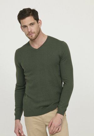 Pull vert en col V avec des manches longues, poignets et ourlet côtelés, design ajusté, fabriqué dans un matériau doux, porté avec un pantalon beige clair.