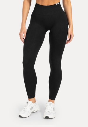 Mujer con leggings negros de cintura alta y zapatillas blancas, de pie con las manos relajadas a los lados sobre un fondo liso.