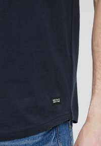 Marineblaues Baumwoll-T-Shirt mit Rundhalsausschnitt und kurzen Ärmeln. Verfügt über ein kleines rechteckiges Etikett am Saum. Getragen mit Jeans.