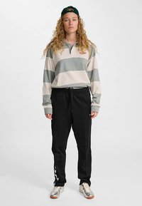 Dolga majica polo s črtami v zeleno-krem barvah, oblečena s črnimi hlačami s stranskimi črtami. Noser z zeleno kapo in supergami.