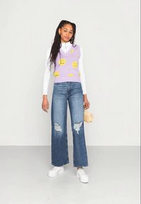 Colete de malha roxo com padrões de rostos sorridentes amarelos, usado sobre um suéter branco de gola alta. Jeans largos azuis com detalhes desgastados e ténis brancos.