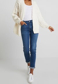 Tjockstickad krämfärgad cardigan över en vit skjorta, ihop med blå jeans och vita sneakers. Cardiganen har ett strukturerat mönster och lös passform.