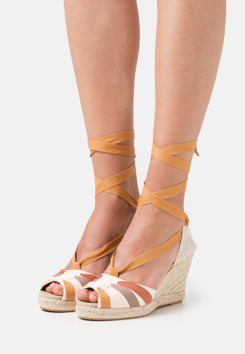 Pieds portant des sandales compensées espadrilles avec des brides multicolores et des rubans couleur marron clair noués autour des jambes inférieures sur fond blanc.
