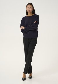 Marineblauer Pullover mit entspannter Passform, Ballonärmeln und runder Halslinie, kombiniert mit schwarzen, maßgeschneiderten Hosen und spitzen schwarzen Schuhen.