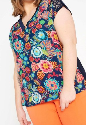 Femme portant un haut bleu marine à manches courtes avec un imprimé floral coloré et des détails en dentelle, associé à un pantalon orange vif.