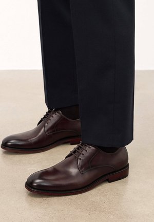 Zapatos de vestir de cuero marrón con punta curva, diseño de cordones y suela roja en contraste, combinados con pantalones oscuros sobre una superficie clara.