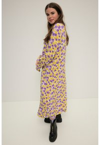 Vestido amarillo con patrón floral púrpura, de mangas largas y botones en los puños. Lleva unos botines negros hasta el tobillo, con una textura de tela suave.