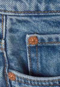 Μπλε τζιν ύφασμα με πορτοκαλί ραφές, που δείχνει μια γωνία τσέπης στερεωμένη με χαλκούς πριτσίνους με ανάγλυφο κείμενο επωνυμίας.