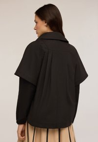 Jachetă neagră cu un guler wide și mâneci scurte, largi. Prezintă un detaliu plisat la spate și manșete elastice la capetele mânecilor.