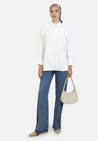 Camicetta bianca con dettagli in pizzo al colletto, abbinata a jeans denim blu a gamba larga. Il modello tiene una borsa beige chiaro.