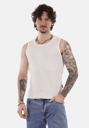 Uomo con capelli ricci che indossa una canottiera bianca e jeans blu, mostra tatuaggi dettagliati su entrambe le braccia, in piedi con una mano in tasca.