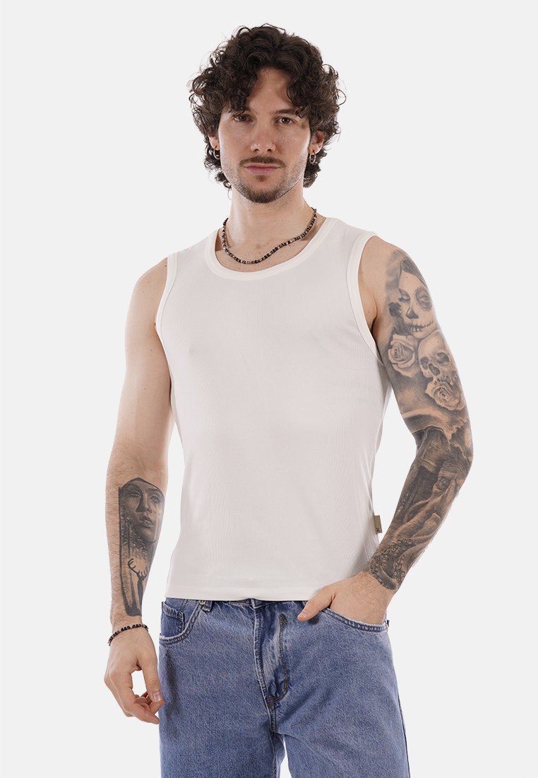 Uomo con capelli ricci che indossa una canottiera bianca e jeans blu, mostra tatuaggi dettagliati su entrambe le braccia, in piedi con una mano in tasca.