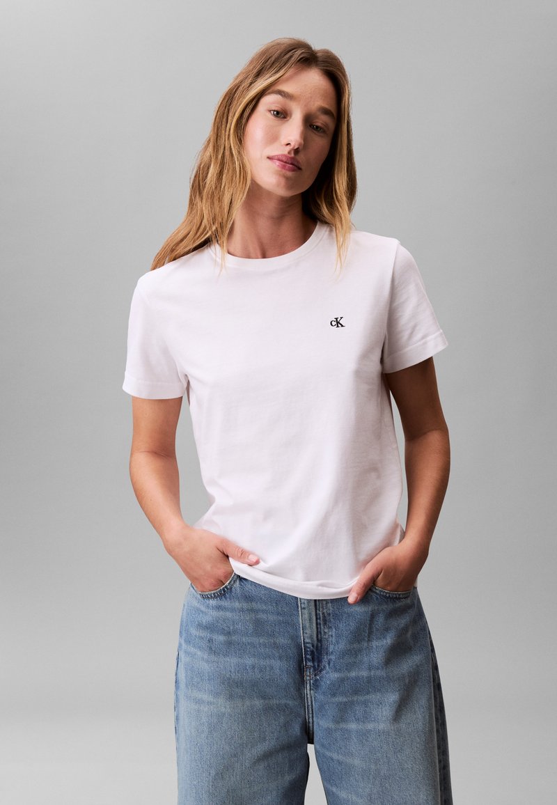 Frau mit langen blonden Haaren, die ein weißes T-Shirt mit kleinem "cK"-Logo und hellblaue Jeans trägt, steht mit den Händen in den Taschen.
