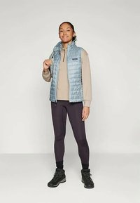 Lys blå puffervest over en beige topp, kombinert med mørke leggings og svarte støvler. Vesten har glidelås og en brystlomme.