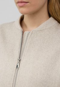 Giacca in pile beige con colletto alto e dettagli con zip argentata, caratterizzata da una texture morbida e un design minimalista. La zip è metallica.