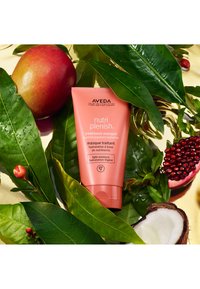 Aveda Nutri Plenish behandlingsmask i en korallfärgad tub, med vit text och logotyp, omgiven av gröna blad, ett rött äpple och kokosnötskal.