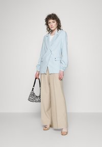 Blazer azul claro com um corte ajustado, mangas bufantes e fecho de um botão; emparelhado com calças largas bejes e uma bolsa com riscas pretas.