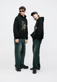 Juodi džemperiai su gobtuvu, puošti grafikiniais dizainais, derinami su tamsiais džinsais ir juodais batais. Priedai apima saulės akinius ir pilką kepurę.