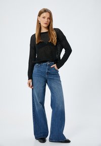 Svart, texturerad långärmad tröja i kombination med högmidjade, vida blå jeans. Modellen poserar med en hand i fickan och visar upp outfiten.