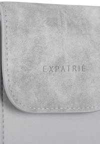 Borsetta in pelle sintetica grigia con una texture liscia, dotata di chiusura a patta, logo "EXPATRIÉ" impresso e dettagli di cucitura visibili.