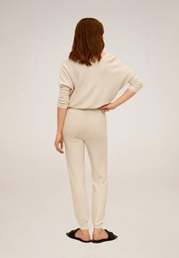 Pull en maille beige et jogging assorti, coupe décontractée, poignets côtelés, design épaules dénudées, porté avec des chaussures noires sans lacets.