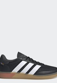 Zwarte sneaker met witte strepen, mesh en leren bovenwerk, zwarte veters en een translucente rubberen zool, voorzien van een Cordura stoffen label.