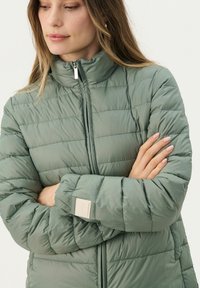 Padded zelena jakna s visokim ovratnikom, horizontalnim šavovima i oznakom na rukavu. Glatka tekstura i lagan dizajn.